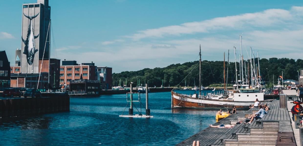 Mennesker soler sig på træplatformen ud til vandet ved Odense havn. Der er blå himmel og lidt skyer. 