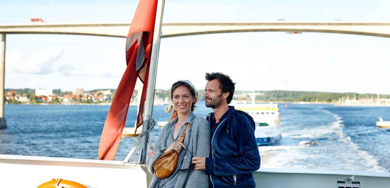 Par står og holder om hinanden på den bagerste del af en færge og nyder udsigten. I baggrunden er Svendborgsund broen. 