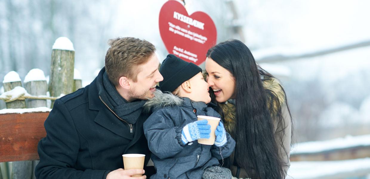 En mand og en kvinde sidder på en 'Kyssebænk' og griner. Imellem dem sidder en lille dreng. Der er sne omkring dem og de er alle klædt godt på.