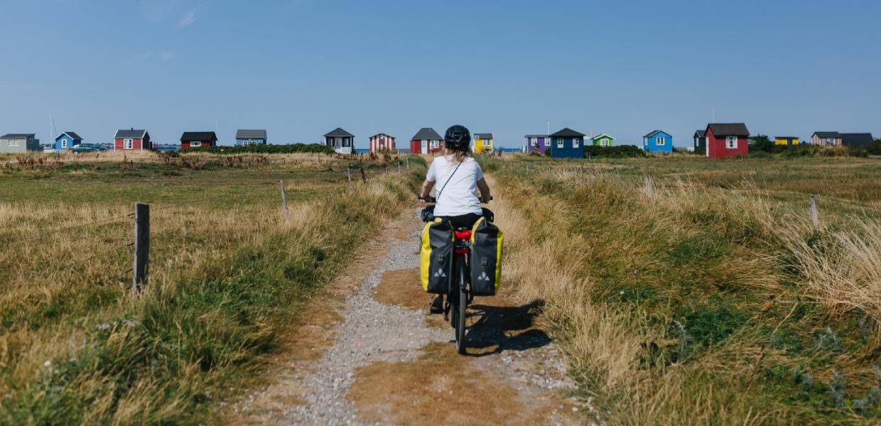 Kvindelig cyklist ses bagfra på vej ud til badehusene ved Vester Strand på Ærø. Det er sommer.