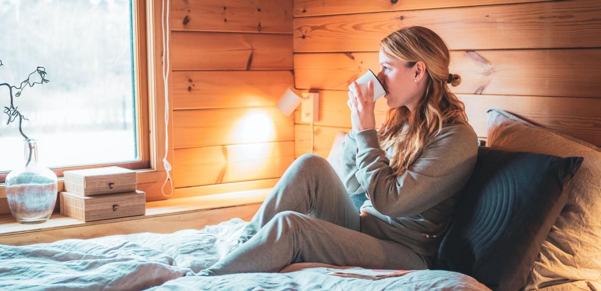 Kvinde sidder afslappet på en seng i en træhytte i behageligt tøj og godt tilbagelænet og drikker af en kop, mens hun kigger ud af vinduet.