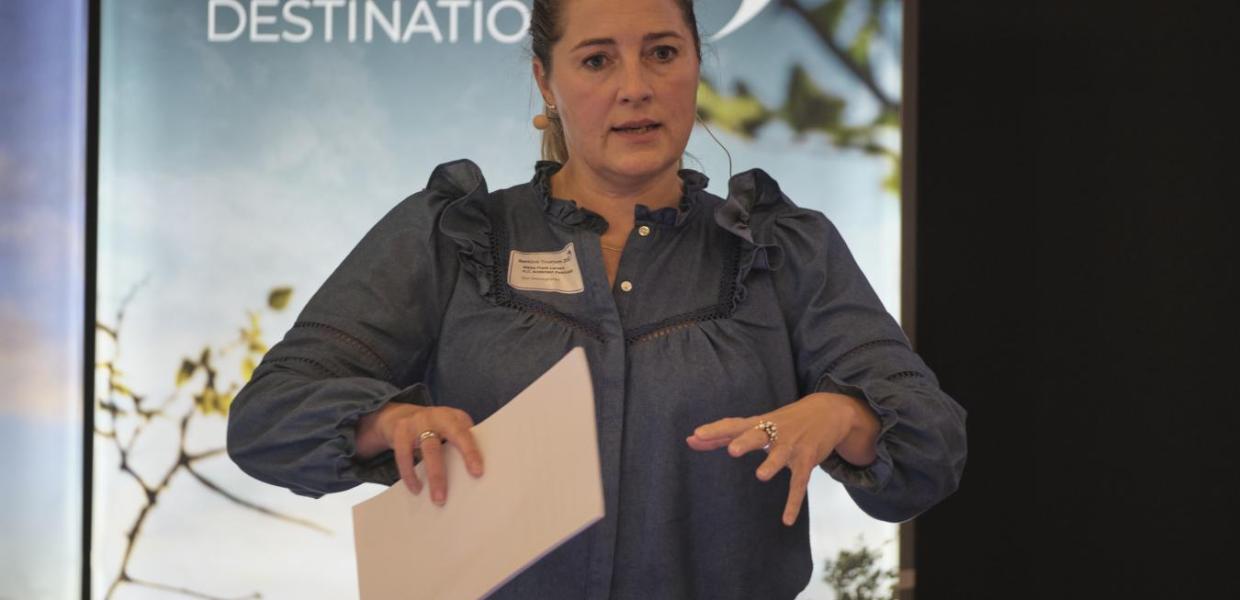 En kvinde med navneskilt og papirer i hånden står og snakker. Bag hende kan man se en planche med 'Destination Fyn', samt deres logo.