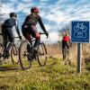 Tre racer-cyklister cykler på en græssti mellem højt græs og træer. I højre side ses et blåt skilt med cykel og navnet Herregårdsruten på.