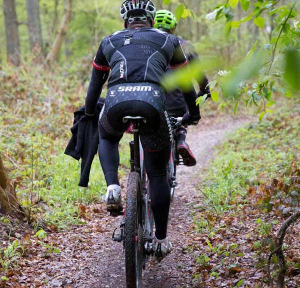 To mountainbikere cykler i skoven, omringet af træer og vildt mos.