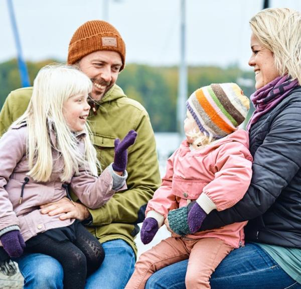 En mand og en dame sidder med hvert sit barn på skødet og i baggrunden er en havn