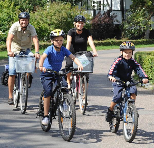 To voksne og to børn cykler af sted på stille asfaltvej med bindingsværkshus i baggrunden. Øverst i højre hjørne ses det røde Bike Friend-logo.