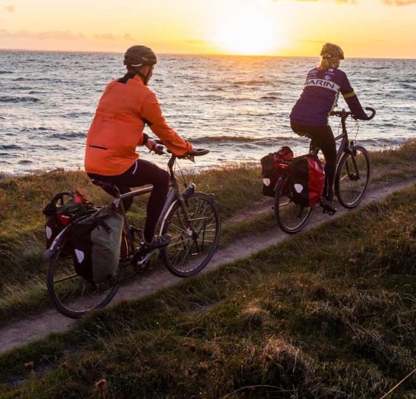 To cyklister cykler på smal grussti lige ud til vandet ved en fynsk strand. I baggrunden ses en smuk solnedgang. 