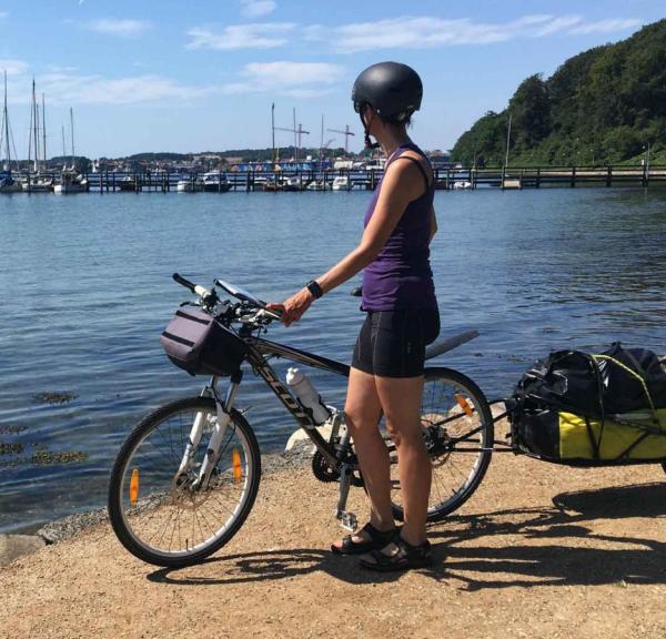En kvinde står på en sti med sin cykel. cykelhjelm og cykeltrailer.  Hun kigger udover havet og havnen der er i baggrunden af billedet.