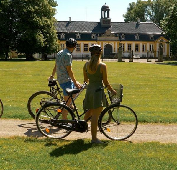 Familie på cykeltur igennem en grøn park, der kigger mod en hvid og gul gård.