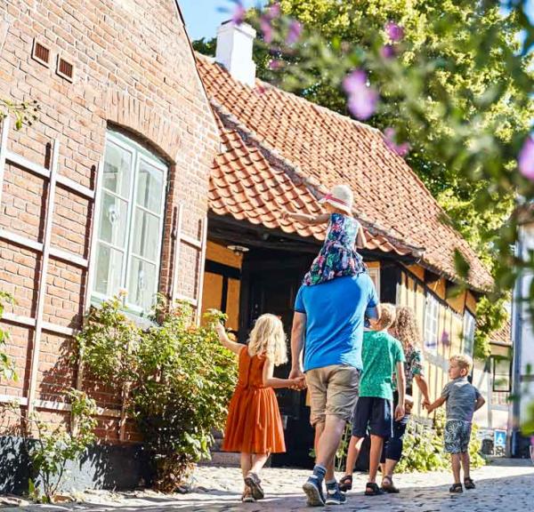 Familie med to voksne og tre børn går ned ad en brostensbelagt gade med bindingsværkshuse og stokroser.