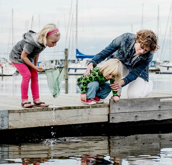 To børn og en voksen fisker med et fiskenet på en havnebro. Skibe i baggrunden. Den voksne holder fast i det mindste barn, der er nede i knæ og læner sig ud over kanten for at se.