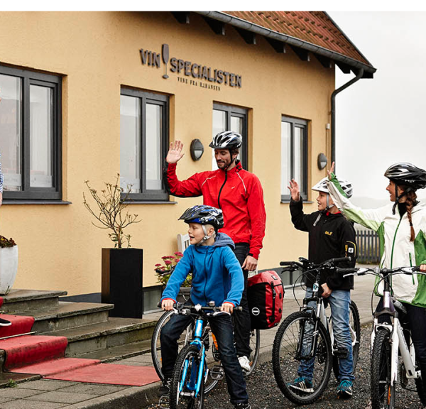 To voksne og to børn på pakkede cykler vinker farvel til en mand, der står på den røde løber på trappen op til VinSpecialisten. Øverst i højre hjørne ses det røde Bike Friend-logo.