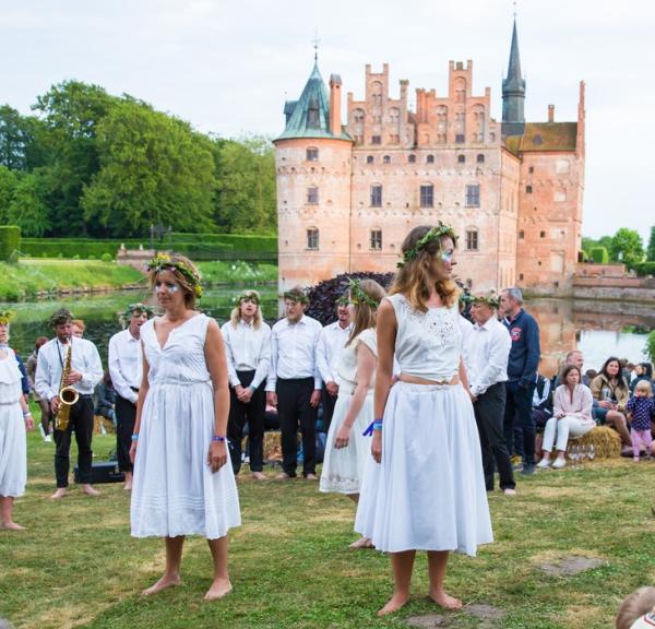 Foran Egeskov slot optræder kvinder i hvide kjoler og blomsterkranse. Omkring dem sidder mennesker og kigger på.