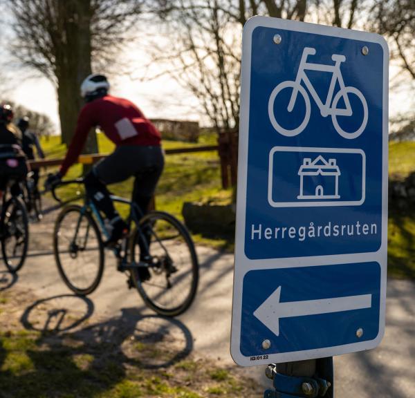 i forgrunden er et blåt skilt med en cykel, pil til venstre og tekst hvor der står Herregårdsruten. I baggrunden tre personer på racercykel ned af en sti der går mellem træer. 