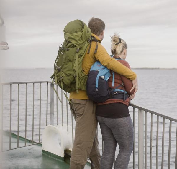 Et par i vandretøj og med rygsække står sammen og kigger ud på havet. De står op af et rækkeværk på bagdækket af en færge.
