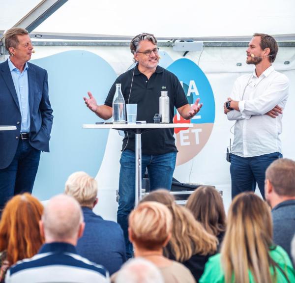 Tre mænd står på en scene og debatterer ved nogle høre caféborde med vandflasker på. I forgrumden ses baghovederne af tilhørerne.