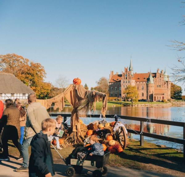 I baggrunden ses i Egeskov Slot omringet af vand. I forgrunden ses mennesker og halloween-pynt i form af græskar, spindelvæv og edderkopper.