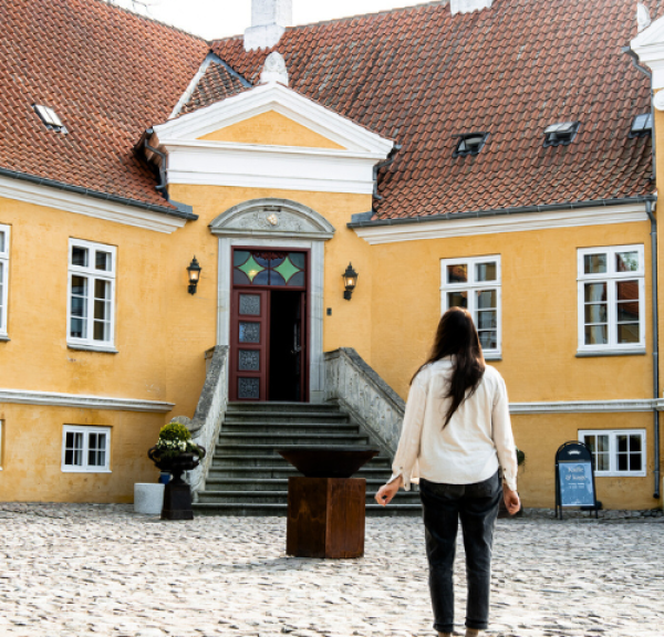 En langhåret kvinde står foran en gul herregård med ryggen til