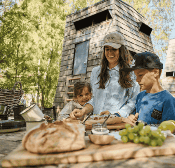 En familie bestående af en mor, en datter og en søn, iført sommertøj, laver mad udendørs foran et shelter