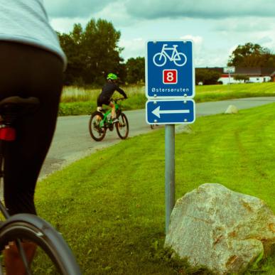 To cyklister kører afsted forbi blåt skilt med hvidt cykelikon, et rødt 8-tal og teksten Østersøruten.