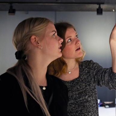 To kvinder står foran en glasmontre. Den ene peger ind i glasmontren. I glasmontren står hvidt porcelæn pyntet med guld detaljer.  