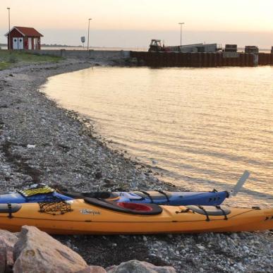 På en strandbred ligger to kajakker i forgrunden. I horisonten af billedet kan man se en  strandspids hvor der står et lille rødt hus foran solnedgangen. 