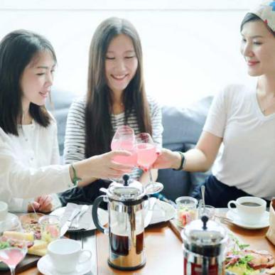 Tre kinesiske piger sidder på række og skåler med vinglas. Bag dem er en lang række af grå sofapuder. Foran dem er et træbord fyldt op med mad, stempel kaffekander og kaffekopper.