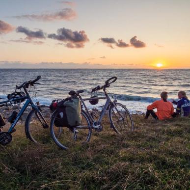 To cykler med oppakning står parkeret i strandkanten. To mennesker sidder ved siden af og ser ud over havet, hvor solen går ned.