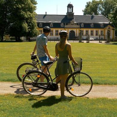 Familie på cykeltur igennem en grøn park, der kigger mod en hvid og gul gård.
