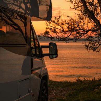 Autocamper i orange solnedgangsbelysning med vand og et stort træ i baggrunden