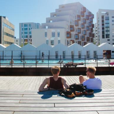 To drenge sidder med ryggen til ved havnen i Odense og kigger ud på havnebadet på en solskinsdag. To piger går en tur foran dem. 
