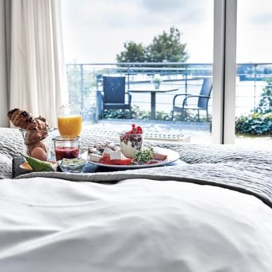 I fodenden af en dobbeltseng med hvid lagner står en bakke med morgenmad. Bagved sengen er der gennem vinduet udsigt til en terrasse der har mark og skov bag sig.