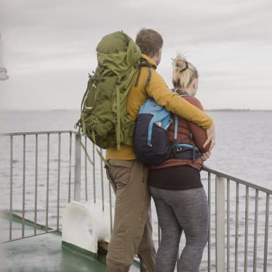 Et par i vandretøj og med rygsække står sammen og kigger ud på havet. De står op af et rækkeværk på bagdækket af en færge.
