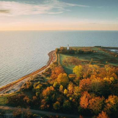 Et dronebillede fra en efterårsdag. Billedet er af en kyststrækning. I venstre side glider havet og himlen sammen i horisonten. Til højre ses en skov ovenfra, og spidsen af land. På spidsen kan man se et lille hvidt fyrtårn. 