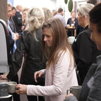 Kvinde i lyserød blazer tapper kaffe i kaffeautomaten på en konference, mens en anden kvinde venter på hendes tur. Mange mennesker i baggrunden.