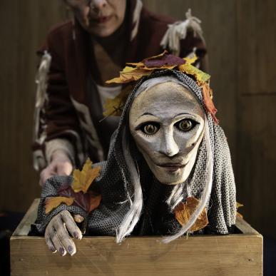 En dukke med hvid maske og gråt hår holdes halvt oppe i en trækasse af en skuespiller i farverigt tøj,