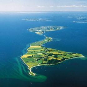 Dronefoto af Avernakø i skarpe, grønne farver med dybblåt vand hele vejen rundt om og horisonten i baggrunden. 