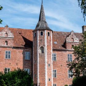 Herregården Harridslevgaard ses gennem flere træer. Gaarden er rød og har i midten et tårn med et helt spidst tag.