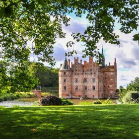Egeskov slot med grønt græs i forgrunden og grønne træer og blå himmel i baggrunden.