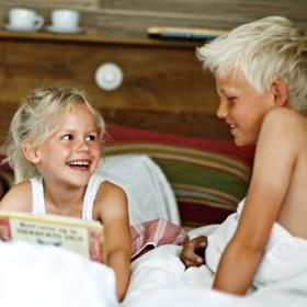 En lyshåret pige og dreng sidder i en seng, kigger på hinanden og griner. Pigen sidder med en bog i hånden.