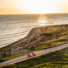 To cyklister set oppefra på vej henover grøn tange med vand til begge sider.