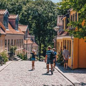Fire personer i sommertøj ses gående ned ad Paaskestræde i Odense. Det er sommer og solen skinner.