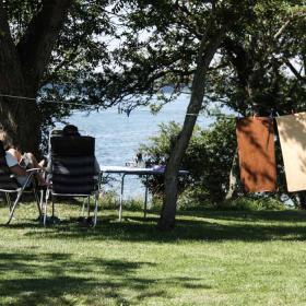 To mennesker sidder på en græsbakke på klapstol og kigger ud på havet. I baggrunden hænger to håndklæder til tørre på en snor og et hvidt telt er slået op.