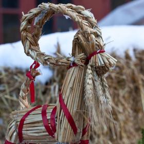 I forgrunden står et rensdyr lavet af halm. Der er bundet et rødt bånd rundt om rensdyret. I baggrunden står en sløret halmballe med sne på. Bag ved halmballen er en rød bygning og en hvid presenning. 