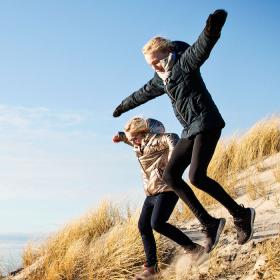 To piger hopper i et klitlandskab i deres overtøj og med blå himmel i baggrunden