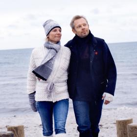 Et par går og holder om hinanden i deres vintertøj mens de kigger fremad og bagved dem er stranden og havet