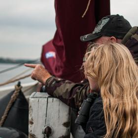 På en båd står en lyshåret pige og en mand i med kasket og camouflage jakke og kigger ud over vandet. Manden peger i den retning de kigger.