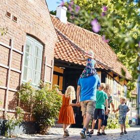 Familie med to voksne og tre børn går ned ad en brostensbelagt gade med bindingsværkshuse og stokroser.