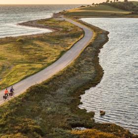 Luftfoto af to cyklister på smal vej ud af tange med vand på begge sider og land i baggrunden.