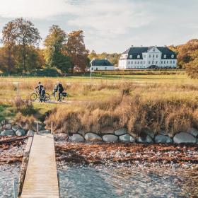 To cyklister gør holdet ved en badebro på strand med dige af store kampesten. I baggrunden en stor, hvid herregård og træer.